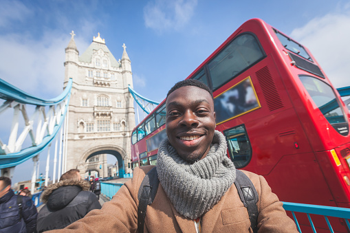 Smiling black man taking selfie in London with Tower Bridge on background. He is holding the phone and looking at camera. Photo taken on a sunny winter day.