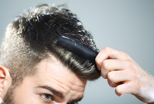 Mid 20's hipster man combing his hair.Using small black comb.He has neat hairstyle with very short hair on the side and one inch length on top,dark brown.Image cut just below his eyes.Gray background.