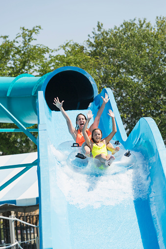 A mother and daughter having fun together at a water park, coming down a giant waterslide on a double innertube, shouting with their arms raised.  They are wearing orange and yellow swimsuits.