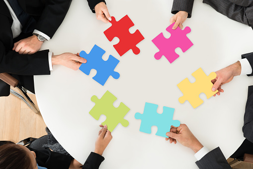 Group Of Businesspeople Holding Multi-colored Jigsaw Puzzle Sitting At The Table