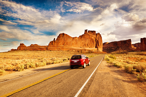 A red car vehicle touring the scenic mountain highways. A popular road trip in the American southwest in Arches National Park in Utah, USA, with its famous rock formation and the dramatic sky in the background. Photographed in horizontal format with copy space.