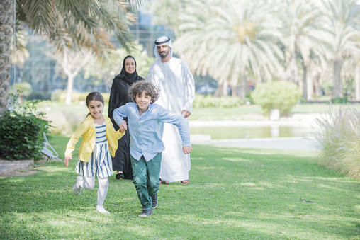 Brother and sister hold hands and run through a park as their mother and father look on from behind. Traditional Arab Emirati family with mother and father wearing thaabe, the woman an abaya and hijab, the man a kandura, ghutra and agal. Dubai, United Arab Emirates.