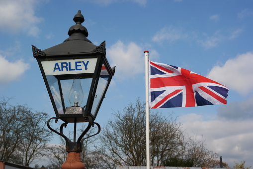 Union Jack flag flying on flagpole in sunlight with the head of a vintage lamp post in front. The lamp post bears the word Arley - the village in Worcestershire, England where the photograph was taken. Both are framed against a blue sky with white clouds