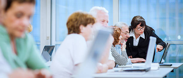 Female Teacher Explaining New Computer Technology to Senior Man Group of adult students in a computer class working behind computers with female teacher explaining, panoramic. Lifelong Learning in Action stock pictures, royalty-free photos & images