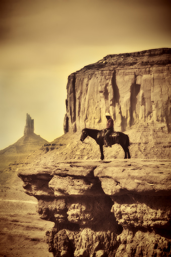 A native American Indian cowboy riding on a horse in the desert landscape in the Monument Valley Tribal Park in Utah, Arizona border, USA. He is on the horse over a cliff with the American southwest plateau in the background. Photographed in vertical format, with desaturated sepia toned effect, with copy space available.