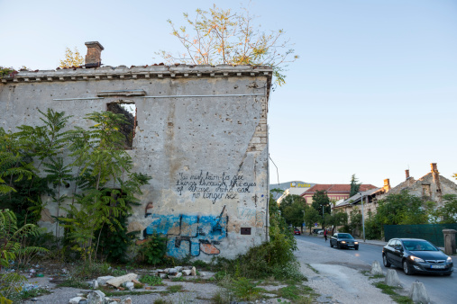 Mostar, Bosnia and Herzegovina - October 3, 2013: Cars drive past a shrapnel and bullet scarred building gutted during the Bosnian War in the early 1990s in Mostar, Bosnia and Herzegovina. A message scrawled in English reads, \