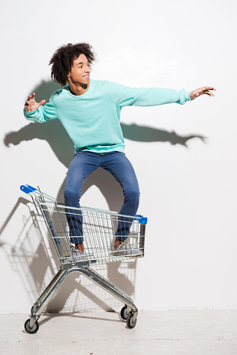 Playful young African man riding in shopping cart against grey background