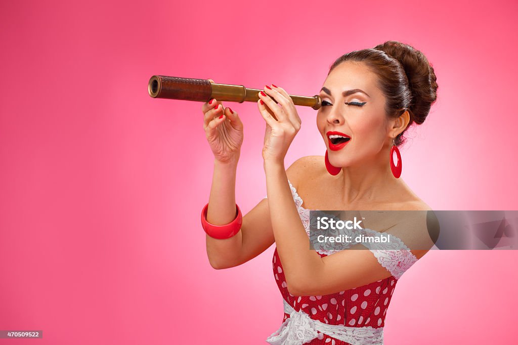 Happy Woman Holding Telescope. Pin-Up Retro style Woman is looking through a telescope and making a surprised expression. She is standing against a pink background. 2015 Stock Photo