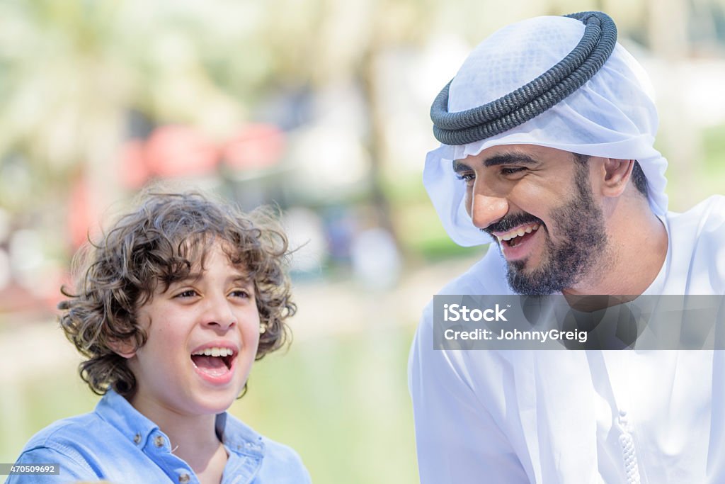 Emirati Arab father laughing with his son Portrait of an Emirati Arab father laughing having fun enjoying time outdoors with his son. He is wearing traditional kandura thobe ghutra and agal. 10-11 Years Stock Photo