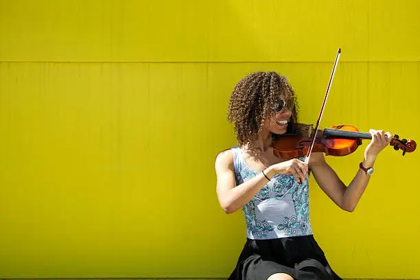 Young African American woman with curly hair playing violin.  Woman is wearing a colorful tank top, black skirt, and sunglasses. Subject is outdoors and isolated on yellow background.