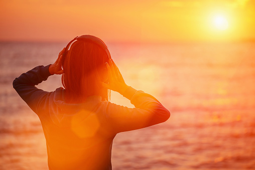 Young woman in headphones listening music and enjoying beautiful sunset over the sea, rear view. Image with sunlight effect