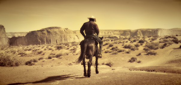 Native American Indian cowboy riding away on saddle horse in the Monument Valley Tribal Park desert landscape, Utah and Arizona border, USA. The American Southwest plateau in a panoramic format, with desaturated sepia tone effect, provides an old, antique, retro revival Wild West impression. Horizontal scenic view with copy space.