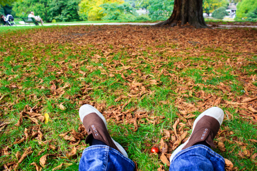 Relaxing in the autumn park - person sitting on the lawn covered with brown fallen leaves with only feet visible in the picture frame.