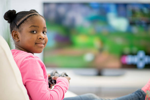 Little girl looking at the camera, and playing video games on a big television in a living room.