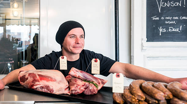 An artisan butcher in a butcher's shop with a sign the reads Venison.  The man is behind the counter and is smiling with a tray of fresh meat in front of him.