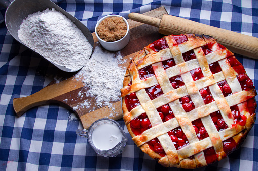 Freshly baked lattice cherry pie just out of the oven placed with ingredients such as flower, brown sugar and cream on a cutting board and blue gingham checked table cloth