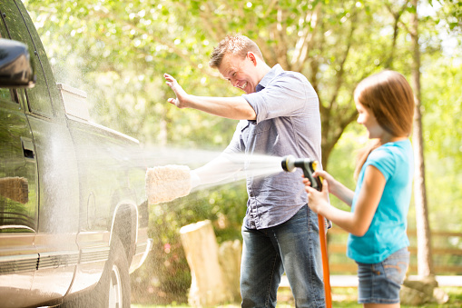 Daddy's little helper! Father and daughter have fun washing the family vehicle together outdoors in the spring or summer season.  They are happy, smiling and laughing as they are enjoying family time.  The little girl uses a water hose sprayer to get the vehicle and dad all wet.  Summer or spring.