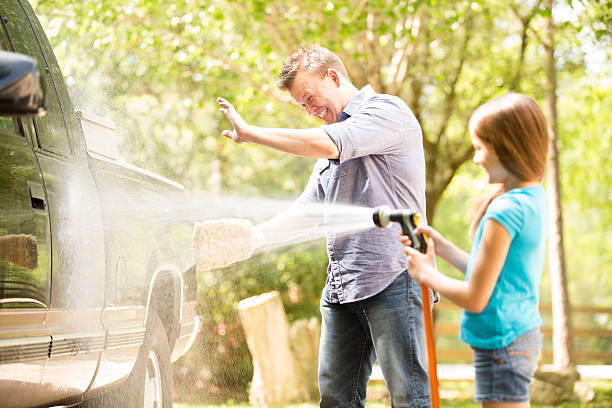 Father and daughter wash the family vehicle together outdoors. Daddy's little helper! Father and daughter have fun washing the family vehicle together outdoors in the spring or summer season.  They are happy, smiling and laughing as they are enjoying family time.  The little girl uses a water hose sprayer to get the vehicle and dad all wet.  Summer or spring. garden hose photos stock pictures, royalty-free photos & images