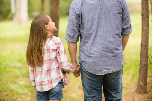 Rear view of father and daughter holding each others hands outdoors in the summer or spring season in their backyard. Girl looks up to father for reassurance. Love and affection for this family of two. The little girl wears a pink plaid shirt and the parent wears a blue shirt.  Sunny nature background.  Special moment for Father's Day.
