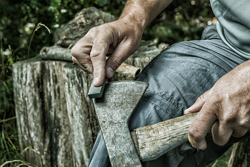 A rustic, rural, gritty adult man's ahnds are sharpening an old, rusty axe manually using a whetstone. Canon 5D Mark II.