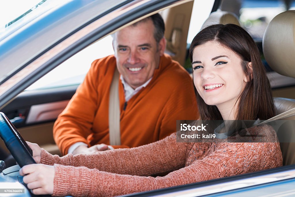 Teenager with father learning to drive A teenager sitting in the driver's seat of a car, looking through the window smiling at the camera.  Her father or a driving instructor is sitting in the passenger seat.  She is learning how to drive. Teenager Stock Photo