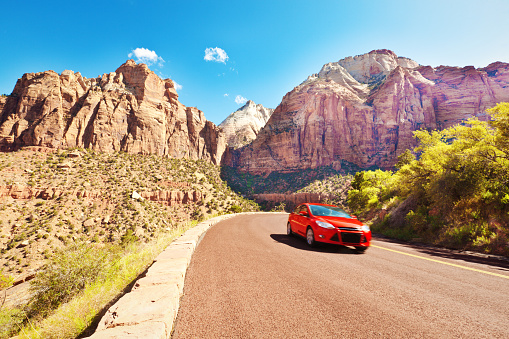 A red car vehicle touring the scenic mountain highways. A popular road trip in the American southwest in Arches National Park in Utah, USA, with its famous rock formation and the dramatic sky in the background. Photographed in horizontal format with copy space.