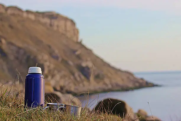 Two silver cups and a blue flask on grass with cliffs and sea in the background.