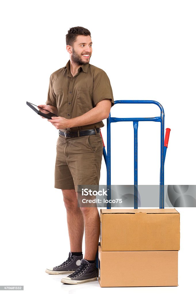 Messenger looking away Smiling courier standing close to a push cart, holding digital tablet and looking back over the shoulder. Full length studio shot isolated on white. Looking Over Shoulder Stock Photo