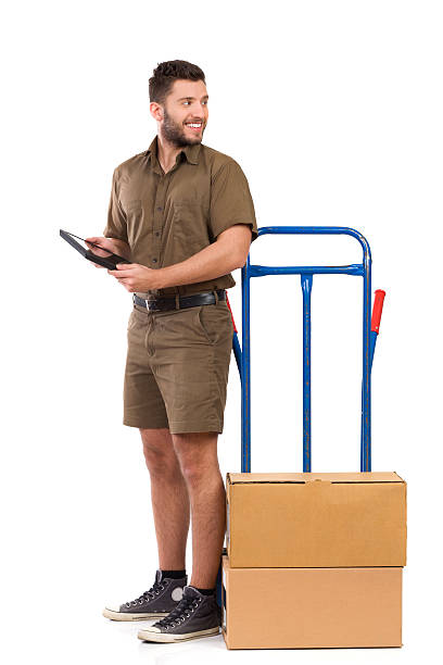 Messenger looking away Smiling courier standing close to a push cart, holding digital tablet and looking back over the shoulder. Full length studio shot isolated on white. mover delivery person box hand truck stock pictures, royalty-free photos & images