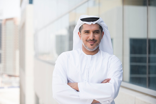 A photo of confident and smiling mid adult arab businessman. Emirati man is wearing tradition clothing. Portrait of middle eastern professional standing arms with crossed outside office building. Dubai, United Arab Emirates, Middle East
