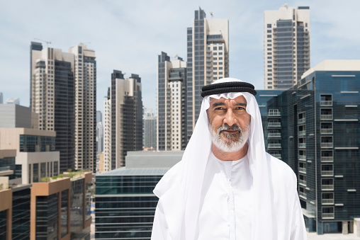 A photo of confident mature arab businessman in traditional clothing of thobe, kandura, ghutra and agal. Portrait of professional is standing with modern skyscrapers in city. Middle Eastern sheikh. Dubai, United Arab Emirates, Middle East