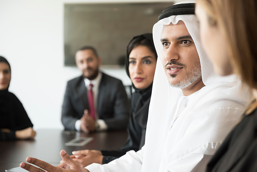 A photo of serious Arab businessman communicating in a meeting. Emirati man is wearing traditional thobe. Middle Eastern professional is discussing while sitting with colleagues, Coworkers are listening to him, in brightly lit office.