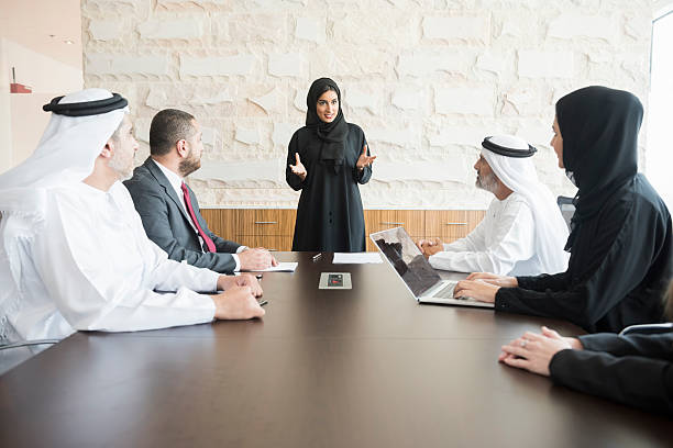 Arab businesswoman giving presentation to colleagues in office A photo of young and smiling Arab businesswoman gesturing while giving presentation. Emirati business people are wearing traditional clothes of those and abaya. Multi-ethnic colleagues are listening to her very carefully. All are in brightly lit office. emirati culture photos stock pictures, royalty-free photos & images