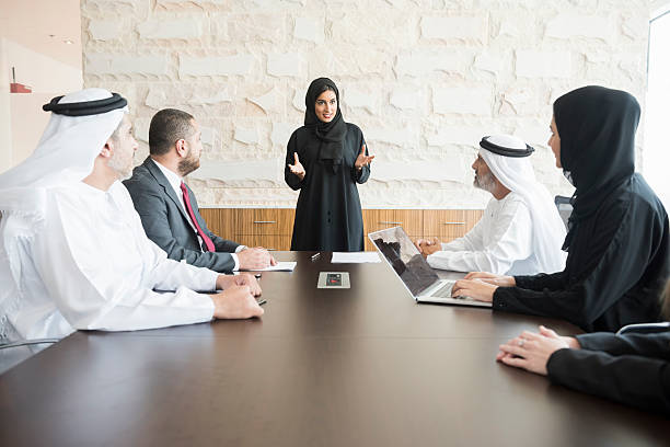 Arab businesswoman giving presentation to colleagues in office A photo of young and smiling Arab businesswoman gesturing while giving presentation. Emirati business people are wearing traditional clothes of those and abaya. Multi-ethnic colleagues are listening to her very carefully. All are in brightly lit office. modest clothing stock pictures, royalty-free photos & images