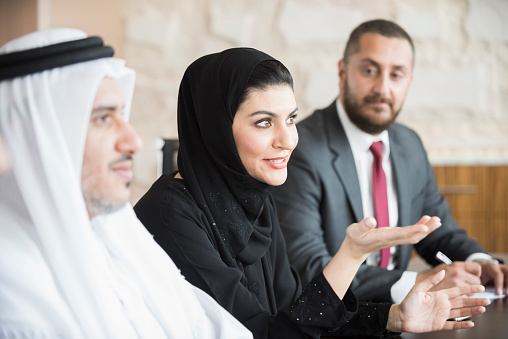 A photo of mature Arab businesswoman talking in a business meeting. Emirati woman is sitting with middle eastern coworkers in board room. Professional woman in traditional abaya and hijab, Dubai, United Arab Emirates, Middle East.