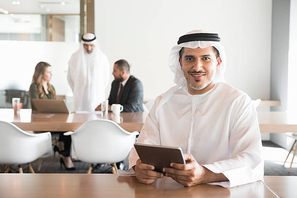 Smiling Arab businessman holding digital tablet in office A photo of confident, smiling Arab man holding digital tablet at desk. Portrait of  professional Emirati businessman in traditional clothing using tablet PC. Focus on him while business colleagues are working in office behind. emirati culture photos stock pictures, royalty-free photos & images