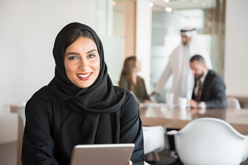 A photo of Emirati businesswoman holding digital tablet at desk. Portrait of middle eastern professional female in traditional abaya using tablet PC. Portrait of young Arab woman at work in modern office in Dubai, United Arab Emirates