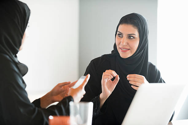 Arab businesswomen in traditional clothes discussing in office A photo of mature Arab businesswoman gesturing while communicating. Emirati woman in traditional abaya at work in office. Middle Eastern female professional is sitting with colleague using mobile phone. modest clothing stock pictures, royalty-free photos & images