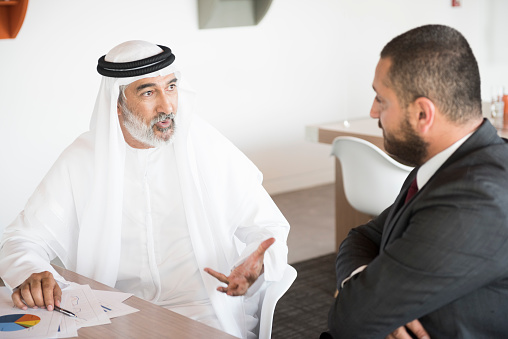 A photo of confident Arab businessman discussing in a meeting with middle eastern business partner. Emirati man wearing traditional clothes. Professional is gesturing while sitting with colleague at conference table, at brightly lit office.