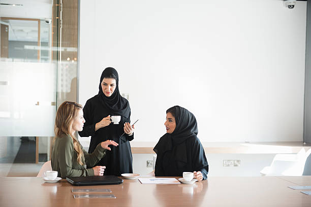 Businesswomen in Middle East office meeting A photo of multi-ethnic businesswomen discussing. Arab Emirati women are in traditional abaya clothes and Caucasian female is in western dress. Professionals are at conference table, in brightly lit modern office discussing business cooperation. Dubai, United Arab Emirates. middle east stock pictures, royalty-free photos & images