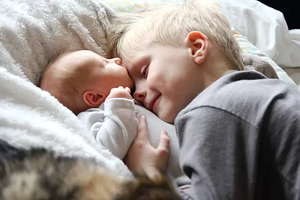 A 5 year old big brother is hugging, smiling, and looking at his newborn baby sister as they sunggle in bed.