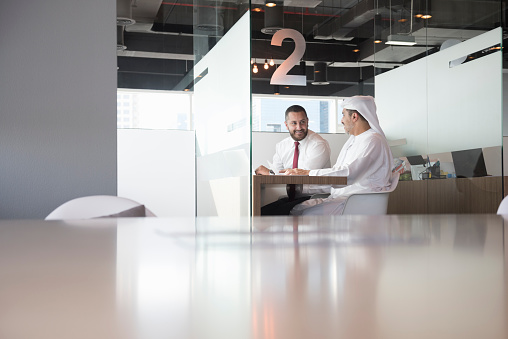 A photo of businessmen smiling together. They are sitting in cubicle. Arab Emirati professionals are sitting at desk. One is wearing formal and other is in traditional middle eastern attire. Desk is in front, at brightly lit office.