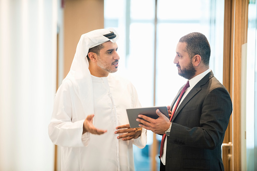 A photo of Emirati  and middle eastern businessmen discussing. Professionals are standing in modern office. One is in suit and other is wearing traditional arab clothes. They are communicating in brightly lit workplace.