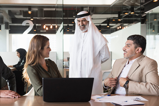 A photo of multi-ethnic arab, middle eastern and Caucasian business people discussing at conference table in modern office. Professionals are with laptop. Two are in formals and other is wearing traditional Emirati attire.