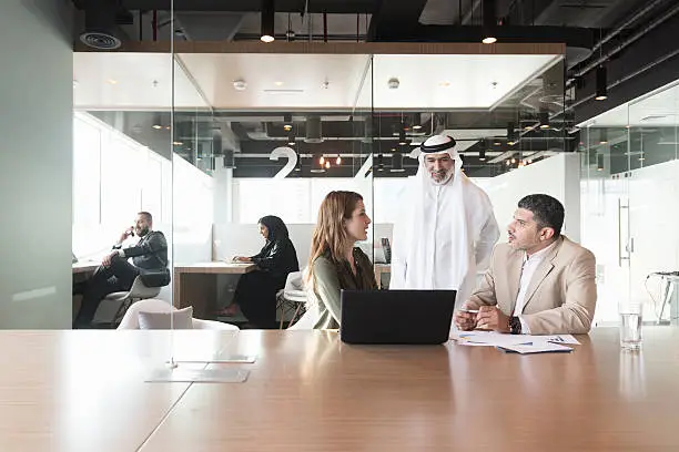 A photo of multi-ethnic Arab, Caucasian and Middle Eastern business people discussing at conference table in modern office. Professionals are with laptop. Some are in formals and others are wearing traditional attire. Dubai, United Arab Emirates.