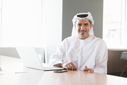 A photo of confident and smiling middle eastern arab businessman is with laptop. Portrait of professional is in traditional clothes. He is sitting at conference table, in a brightly lit office. Desk is found near windows. Dubai, United Arab Emirates.