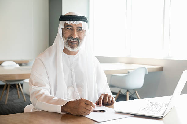 Arab businessman confident and smiling in office A photo of confident and smiling Emirati businessman with laptop. Portrait of professional is in traditional thobe. He is sitting at conference table, in a brightly lit office. Desk is found near windows. Dubai, United Arab Emirates, Middle East emirati culture photos stock pictures, royalty-free photos & images