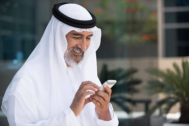 Arab man using mobile phone outside office A photo of smiling arab businessman using mobile phone. Emirati man is standing outside office building and text messaging on smart phone. Happy middle eastern professional is wearing traditional clothes. Dubai, United Arab Emirates, Middle East kaffiyeh stock pictures, royalty-free photos & images