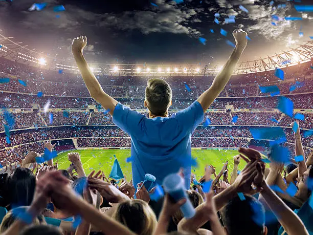 On the foreground a group of cheering fans watch a sport championship on stadium. One man stands with his hands up to the sky. People are dressed in blue colors. A long-range shot of a stadium field, floodlights and seating. A green field, with painted white lines, is visible in the foreground. In the background are diffuse out-of-focus stadium seats. Large, bright floodlights are in the top-left and top-right corners of the image.