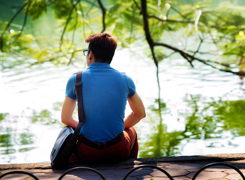Rear view of vietnamese student sitting alone at Ho Hoan Kiem park in Hanoi Vietnam. He is looking left pensively. Willow trees are in the background, touching the lake's water.
